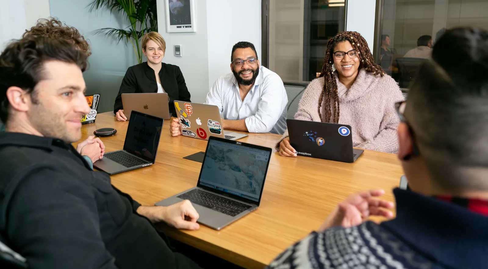 A group of people sit around a table, in a conference room. In front of each person is a laptop and some of the laptops have stickers placed on the back. Image Source: Mapbox via Unsplash